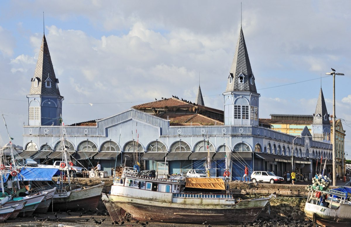 Mercado de Ferro do Ver-o-Peso, com suas torres pontiagudas, e barcos de pesca atracados na doca em frente, sob céu azul com nuvens.