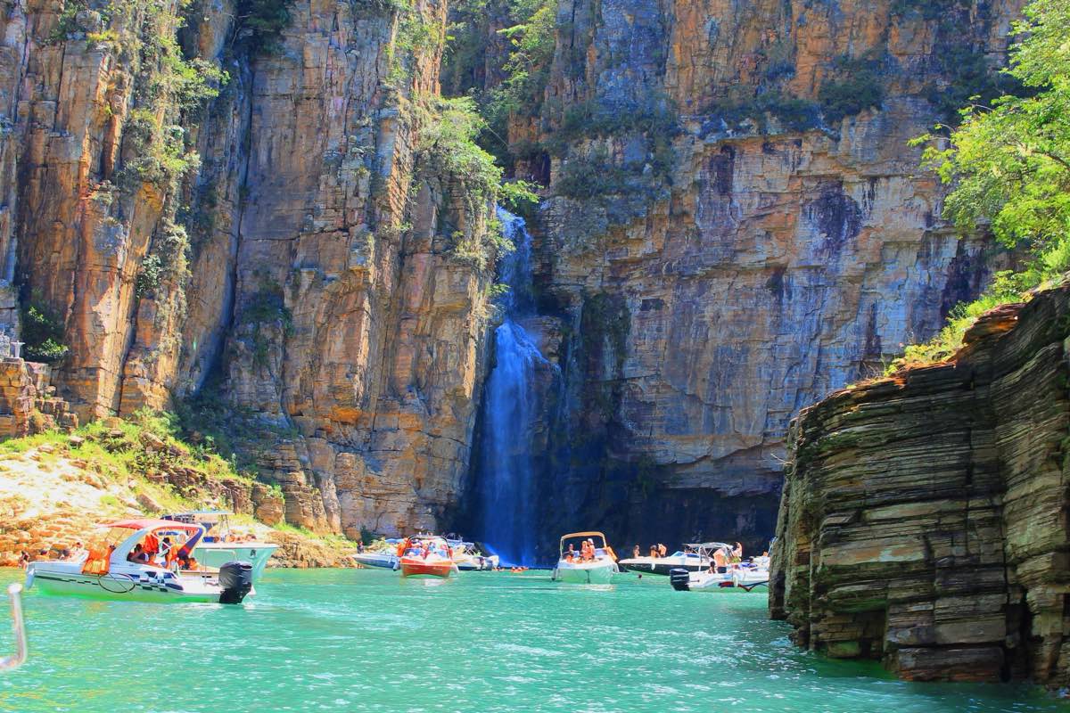 Cachoeira despencando de um paredão de cânion, com lanchas de passeio na água verde-azulada do Lago de Furnas, em Capitólio.