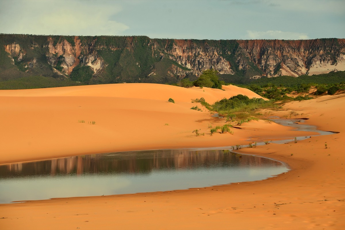Dunas alaranjadas do Jalapão com uma lagoa rasa em primeiro plano refletindo o paredão da Serra do Espírito Santo ao fundo.