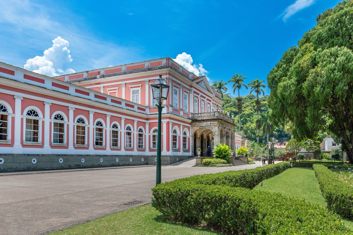 Fachada rosa e branca do Museu Imperial, antigo palácio de verão de Dom Pedro II, com cercas vivas aparadas, palmeiras e céu azul com nuvens.